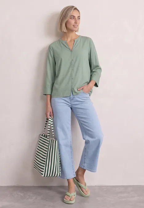 Blouse met 3/4-mouwen en split in de hals eucalyptus green Vrouw met kort blond haar, groen shirt, jeans, groene sandalen en gestreepte tas voor een eenvoudige achtergrond.