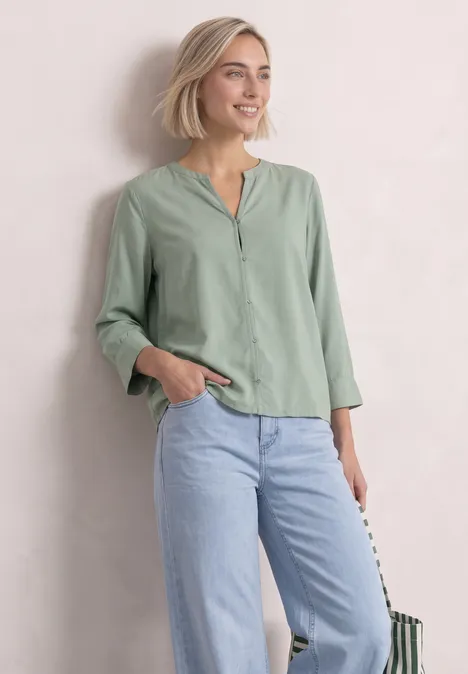 Blouse met 3/4-mouwen en split in de hals eucalyptus green Vrouw met kort blond haar in lichtgroene blouse met V-hals en lichte jeans, glimlachend voor een lichte achtergrond.