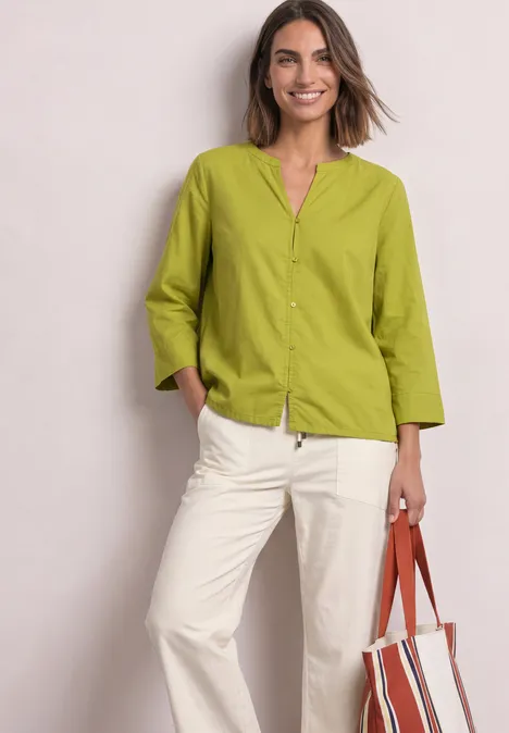 Blouse met 3/4-mouwen en split in de hals apple lime Vrouw in lichtgroene blouse en witte broek glimlacht en houdt een rood-witte tas vast.
