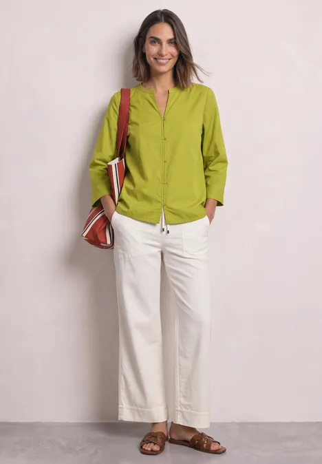 Blouse met 3/4-mouwen en split in de hals apple lime Een lachende vrouw poseert voor een lichte muur, met een groen overhemd, witte broek, bruine sandalen en tas.
