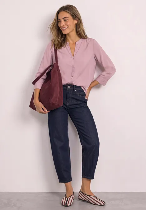 Femme souriante appuyée contre un mur, chemise rose, jean foncé, ballerines à rayures et grand sac bordeaux.