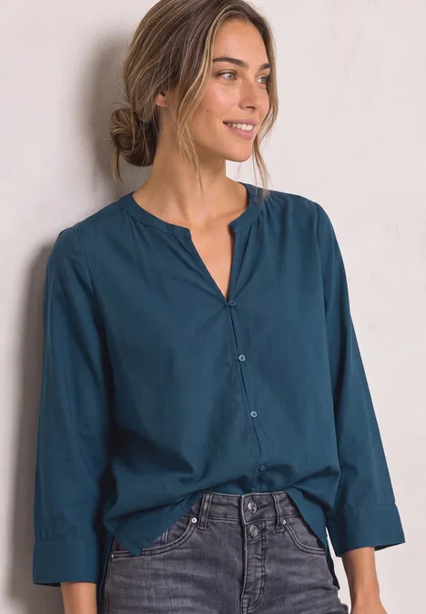 Femme souriante en chemisier bleu à manches longues et cheveux attachés, fond clair.