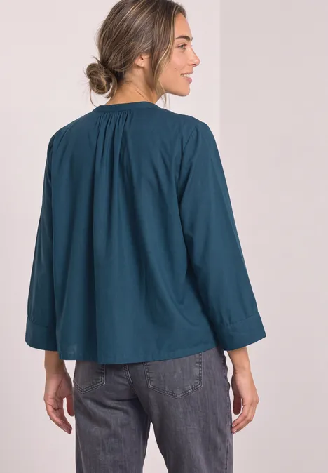 Femme de dos portant un chemisier bleu-canard à manches larges et un jean.