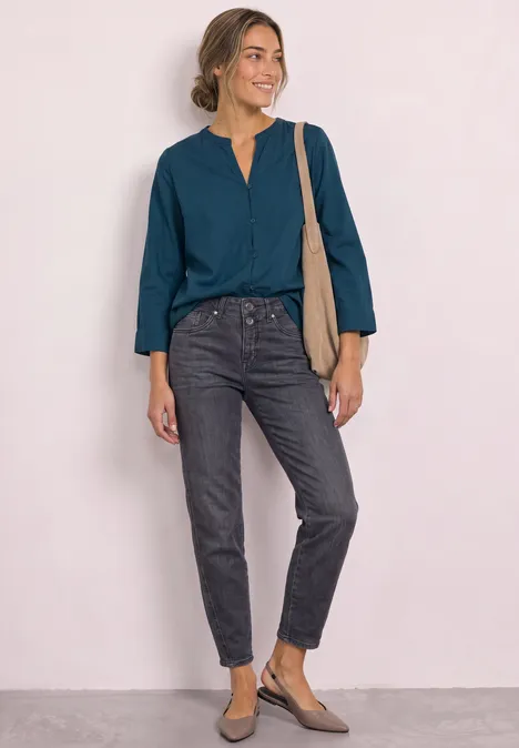 Femme portant un chemisier bleu pétrole, jean noir, chaussures plates et sac bandoulière beige, souriante debout.