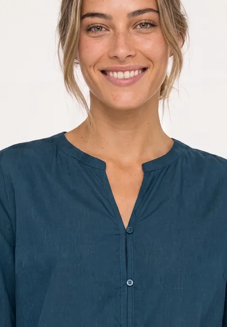 Une femme souriante en chemisier bleu foncé sans col, fond clair.