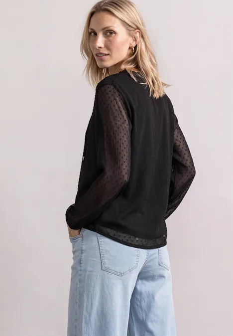 Frau in schwarzem Top mit transparent gepunkteten Ärmeln und hellblauen, weiten Jeans blickt über die Schulter.
