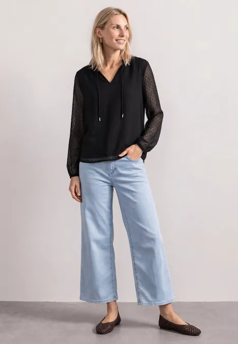 Frau trägt schwarze, leicht transparente Bluse mit Kapuze, hellblaue, weite Jeans und flache Schuhe.