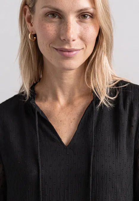 Eine lächelnde blonde Frau mit schwarzem Oberteil und Sommersprossen vor hellem Hintergrund.