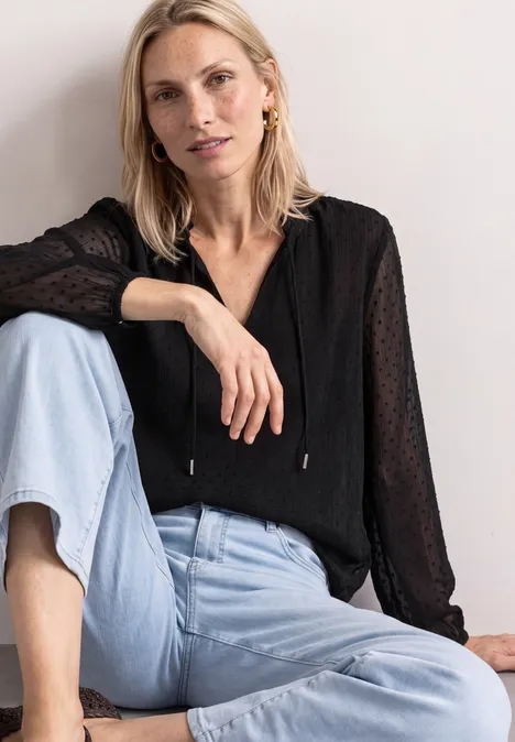 Eine blonde Frau sitzt entspannt, trägt eine schwarze Bluse und hellblaue Jeans.