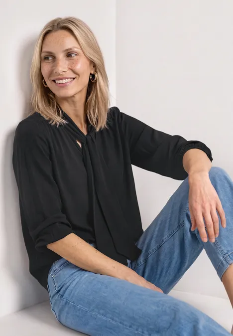 Langarm Bluse mit Schleifendetail Black Eine Person sitzt mit schwarzer Bluse und blauen Jeans an einer hellen Wand.