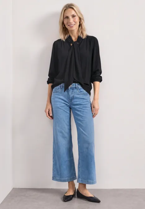 Langarm Bluse mit Schleifendetail Black Frau trägt schwarze Bluse, weite hellblaue Culotte-Jeans und schwarze Ballerinas vor weißem Hintergrund.