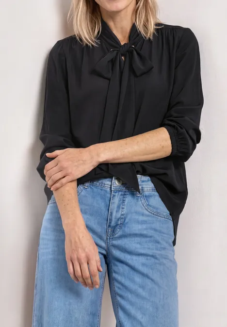 Langarm Bluse mit Schleifendetail Black Frau trägt schwarze Bluse mit Schleife am Kragen und blaue Jeans, Arme vor dem Bauch verschränkt.