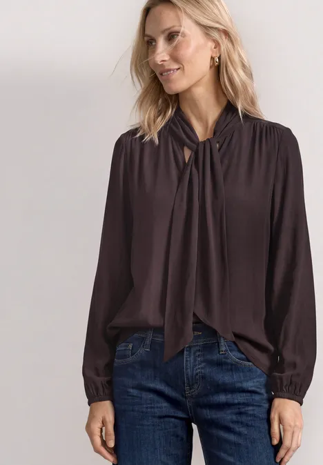 Blouse met lange mouwen en strikdetail ruby vine Frau trägt dunkelbraune Bluse mit Knoten am Kragen und Jeans.