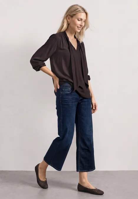 Blouse met lange mouwen en strikdetail ruby vine Frau trägt schwarze Bluse, weite Jeans und flache Mokassins, steht seitlich vor neutralem Hintergrund.