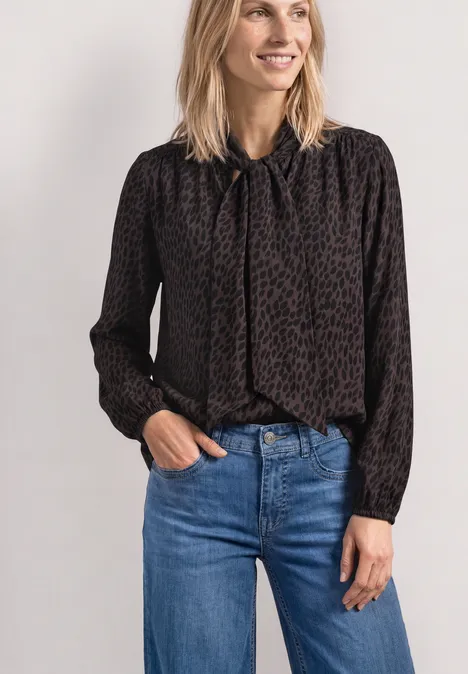 Langarm Bluse mit Schleifendetail molten earth Eine Frau trägt eine schwarze, gemusterte Bluse mit Schleife am Hals und blaue Jeans.