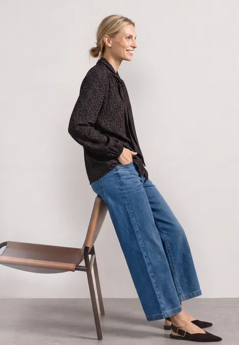 Langarm Bluse mit Schleifendetail molten earth Seitlich abgebildete Person mit schwarzem Hemd, weitem Jeans, schwarzen Loafers lehnt an einem Holzstuhl.
