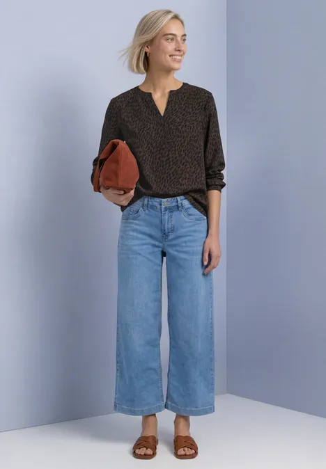 Langarm Bluse mit Split Neck und Print molten earth Frau vor neutralem Hintergrund in schwarzer Bluse mit Tupfen, weitem Jeans, braunen Sandalen und roter Clutch.