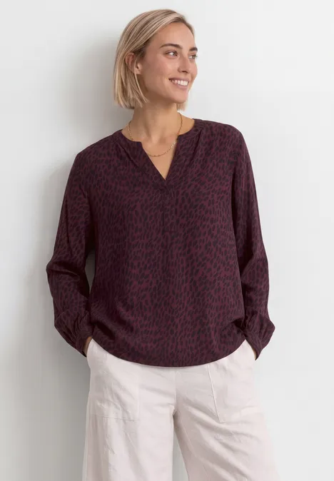Blouse met lange mouwen, split in de hals en print rustic rouge Een glimlachende vrouw met kort blond haar, gekleed in een bordeauxblouse met print en een lichte pantalon.