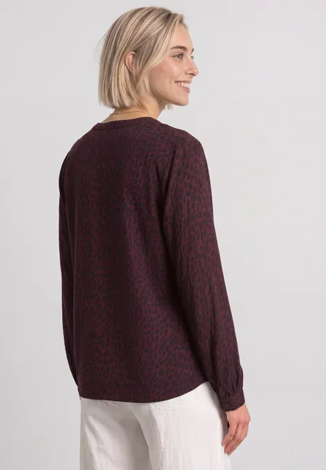 Blouse met lange mouwen, split in de hals en print rustic rouge Vrouw met kort blond haar draagt een donkerrood, gemêleerd shirt met lange mouwen en een witte broek.