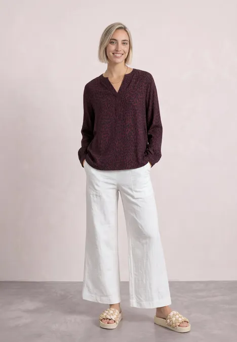 Blouse met lange mouwen, split in de hals en print rustic rouge Vrouw in donkerroze dessinblouse, witte broek en sandalen tegen lichte achtergrond.