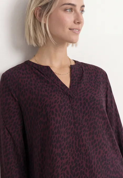 Blouse met lange mouwen, split in de hals en print rustic rouge Vrouw met kort blond haar draagt soepel vallende donkerrode blouse met zwarte dierenprint en V-hals.