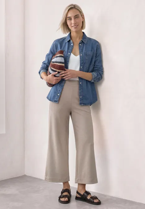 Overhemd met lange mouwen en borstzakken Medium Blue Soft washed Frau in lässigem Outfit aus Jeanshemd, weißem Top und beigen Hosen hält einen gestreiften Ball und lächelt.