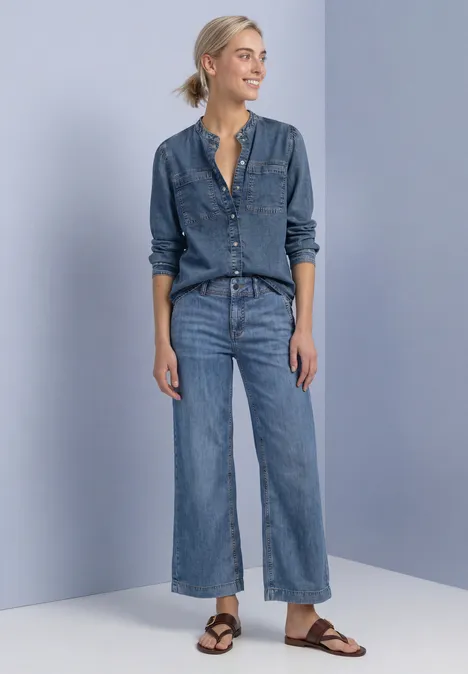 Blouse met lange mouwen en borstzakken Medium Blue Soft washed Eine lächelnde Frau trägt Jeanshemd, weite Jeans und braune Sandalen vor hellgrauer Wand.