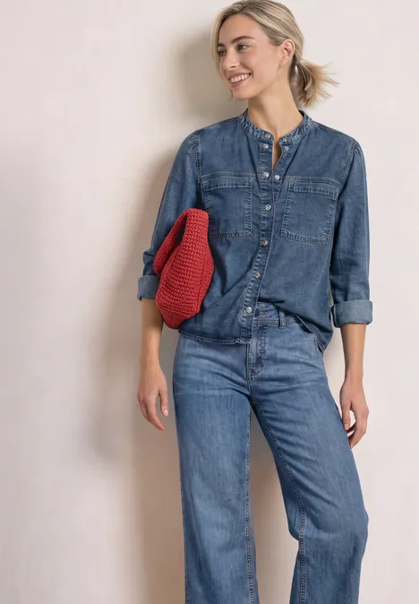 Blouse met lange mouwen en borstzakken Medium Blue Soft washed Eine Frau in blauem Jeans-Outfit lehnt lächelnd an einer Wand und hält einen roten Pullover.