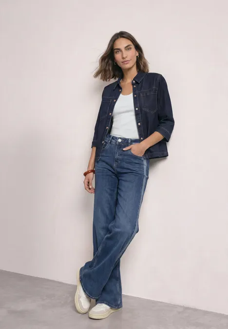 Denim overhemd in rinsed-look Rinse Soft Washed Frau in Jeansjacke, weißem Top und weit geschnittenen Jeans lehnt lässig an einer hellen Wand.