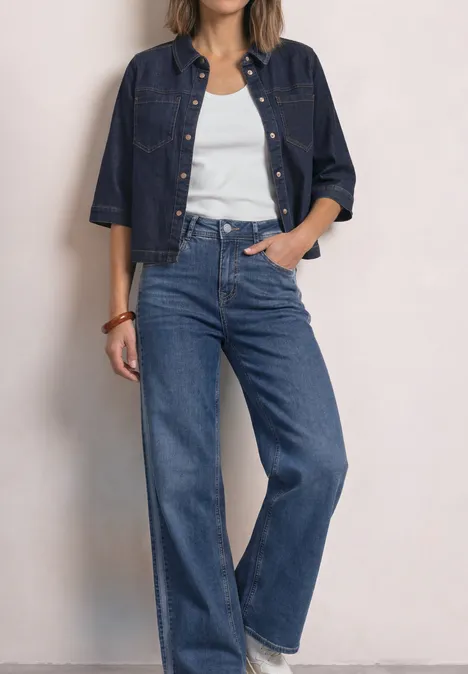 Denim overhemd in rinsed-look Rinse Soft Washed Frau trägt weißes Tanktop, weite blaue High-Waist-Jeans und dunkelblaue Jeansjacke.