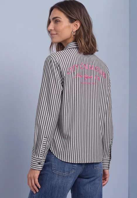 Eine Frau in schwarz-weiß gestreiftem Hemd und Jeans blickt über die Schulter, Schriftzug auf dem Rücken.