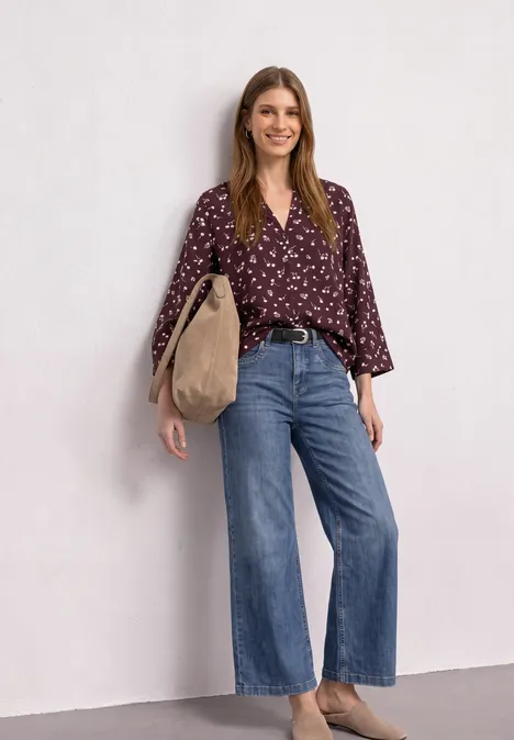 Eine lächelnde Frau trägt eine weinrote Blumenbluse, weite Jeans und eine große beige Tasche über der Schulter.