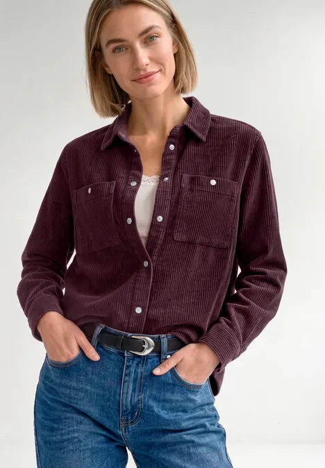 Vrouw in pruimkleurig corduroy overhemd, wit topje en jeans, voor een lichte achtergrond.