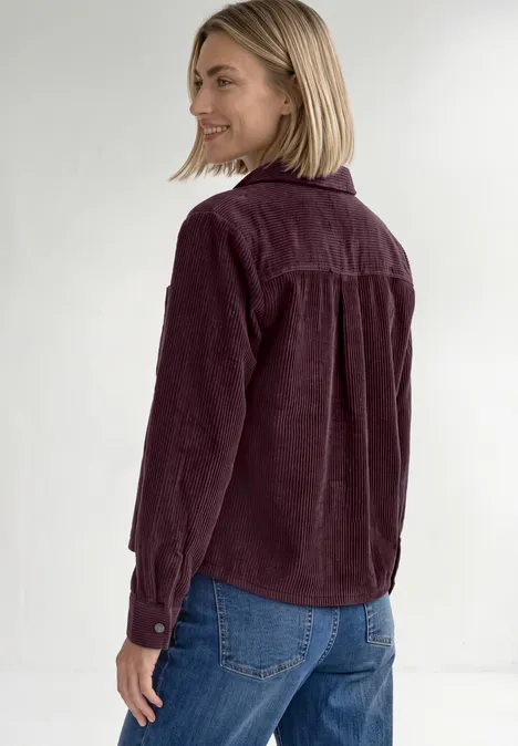 Blonde vrouw draagt auberginekleurig corduroy overhemd en blauwe jeans, kijkt lachend opzij.