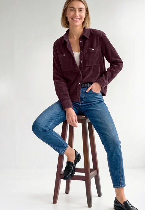 Vrolijke vrouw zit op een kruk, draagt een bordeaux corduroy jasje, witte top, spijkerbroek en zwarte loafers.