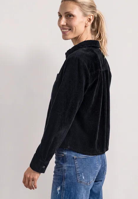 Een glimlachende vrouw in een zwarte ribfluwelen jas en blauwe jeans, zijwaarts poseerend.