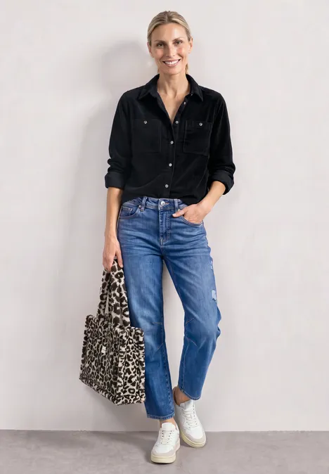 Vrouw in zwart hemd, blauwe jeans en witte sneakers, met panterprinttas, leunend tegen een witte muur.