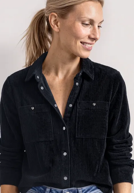 Vrouw met blond haar in een staart draagt een zwarte corduroy blouse en lacht opzij.