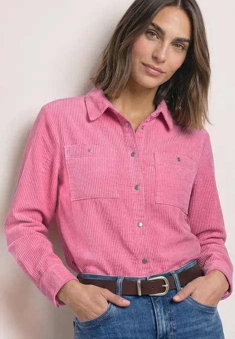 Vrouw met roze blouse en spijkerbroek, handen in zakken, glimlachend voor een lichte achtergrond.