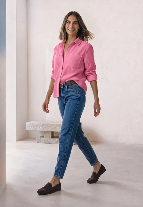 Een vrouw in een roze blouse en blauwe jeans loopt elegant door een lichte, minimalistische ruimte.