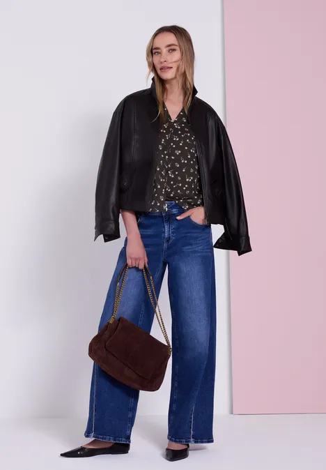 Langarm Chiffon-Bluse mit Split Neck black coffee Frau mit schwarzer Lederjacke über den Schultern, gemusterter Bluse, weitem blauen Jeans und brauner Tasche vor Wand.