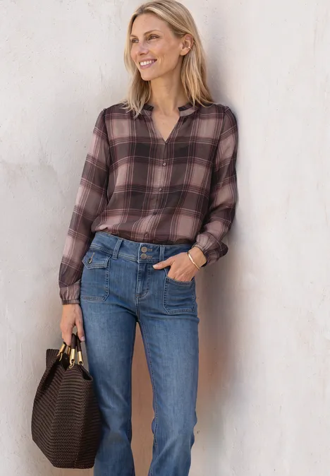 Eine lächelnde Frau in karierter Bluse und blauen Jeans lehnt entspannt an einer Wand und hält eine braune Tasche.