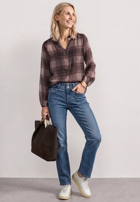 Frau mit kariertem Hemd, blauen Jeans, weißen Sneakern und Tasche, steht lächelnd.