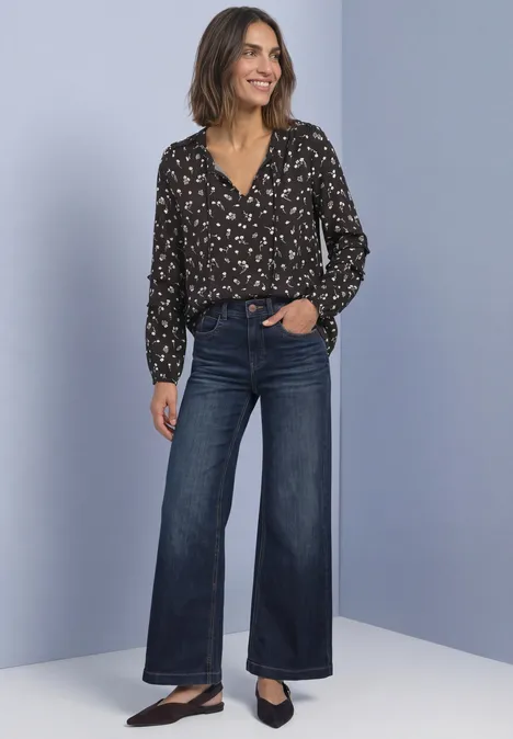 Frau in schwarzer, gemusterter Bluse und weiten, dunklen Jeans, lächelnd vor hellem Hintergrund.