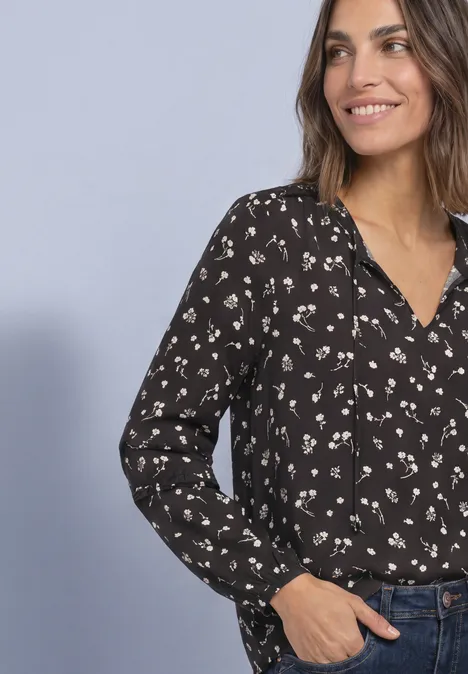 Eine lächelnde Frau trägt eine schwarze Bluse mit hellem Blumenmuster und hat die Hand in der Jeans-Tasche.