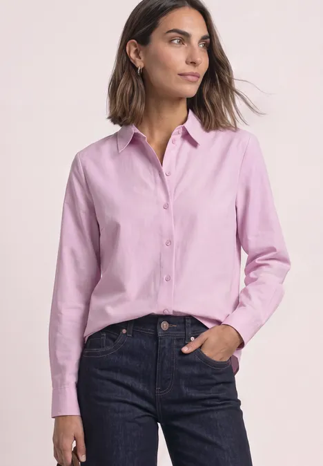 Vrouw in een roze blouse en donkere spijkerbroek, hand in de zak.