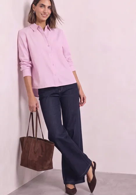 Vrouw in roze blouse, wijde jeans en bruine tas, nonchalant leunend tegen een muur.