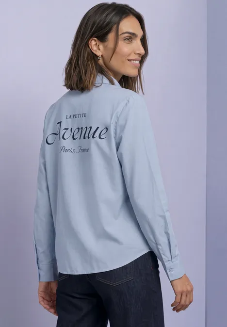Femme en chemise bleu clair avec inscription 'Avenue Paris New' dans le dos, regard par-dessus l’épaule, souriante.