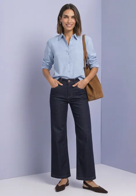 Femme en chemise bleue, jean large et sac marron devant un fond violet pâle.