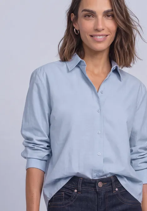 Femme en chemisier bleu clair ample, manches retroussées, semi rentrée, qui sourit doucement.